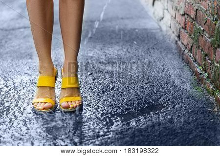 Female feet standing in the rain on wet pavement near a brick wall. Yellow sandals on the feet of a young girl. Water splashes and drops on his feet.