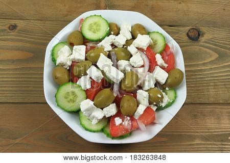 Greek salad from fresh organic vegetables: chopped red onion tomatoes cut on wedges sliced seedless cucumber; and stuffed green colossal olives cubes goat Feta Cheese in white dish on wooden table