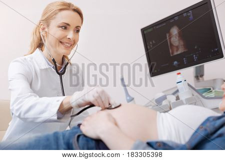 Checking fetus viability. Skilled caring female gynecologist sitting in the sonography cabinet while examining pregnant belly and using stethoscope for detecting heat beating