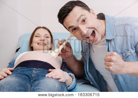 Enjoying first sound of my child. Amused happy delighted husband sitting in the clinic and using medical equipment while listening to the fetus heart beating in the pregnant belly