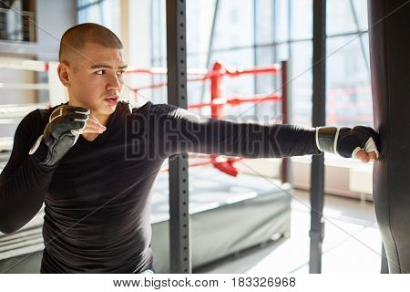 Side view portrait of young muscular sports man hitting punching bag during boxing practice in fight club, training speed and strength