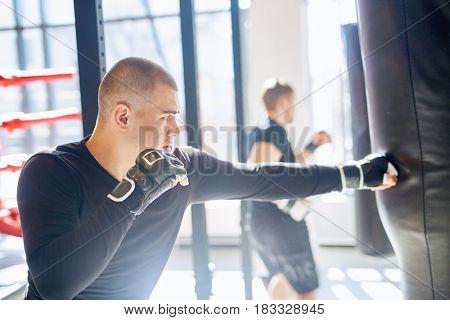 Side view portrait of young muscular boxer hitting punching bag in sunlit fighting clubpracticing speed and strength