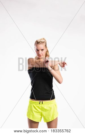 Attractive young fitness woman in black tank top and neon yellow shorts stretching arm. Studio shot on gray background.
