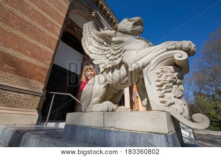 three years old blonde child saluting leaning on winged lion statue outside Palace of Velazquez (from year 1883 by architect Ricardo Velazquez Bosco) in public park El Retiro Madrid Spain