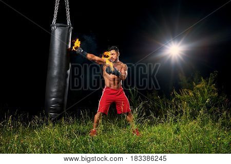 Shirtless muscular male boxer training outdoors at night exercising on a punching bag with his boxing gloves burning with fire copyspace fitness lifestyle forceful powerful confidence athlete.