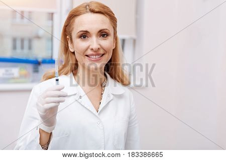Precautionary measures. Attractive female keeping smile on her face holding syringe in right hand looking straight at camera