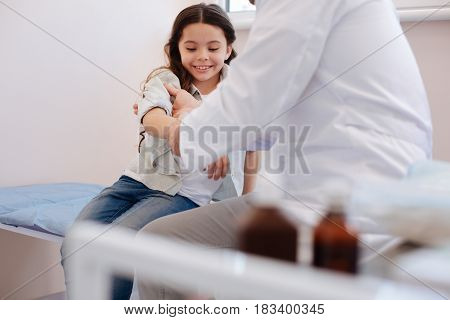 My arm hurts. Nice positive cheerful girl sitting on a medical med and holding up her arm while being examined by a rheumatologist