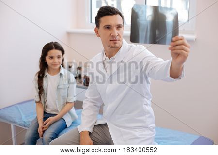 Professional rheumatologist. Serious professional handsome doctor holding an X ray photo and examining it while sitting near his patient