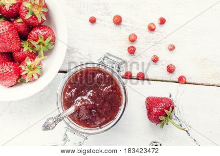 Homemade delicious strawberry jam in a glass jar and fresh strawberries on white rusric wooden background. Top view.