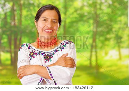 Closeup beautiful hispanic woman wearing traditional andean white blouse with colorful decoration around neck, matching red necklace and ear, posing happily for camera holding out palm of hand, garden background.