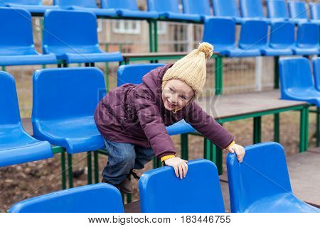 Little girl in a violet coat and a beige woolen hat sitting in a blue seat on a stand.