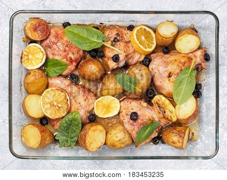 Top view of chicken thighs with potatoes, lemon and black olives, cooked in oven on gray concrete background. Baked chicken leg quarter in heat-proof glass.