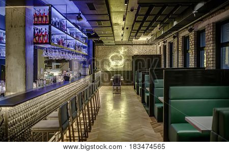 Stylish glowing cafe in a loft style with brick walls and concrete column. There is a bar rack with chairs, shelves with bottles and glasses, bar equipment, sofas with tables, luminous burger on wall.