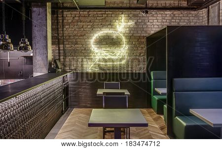 Cafe in a loft style with brick walls, concrete column and a parquet on the floor. There is a textured bar rack, fancy hanging lamps, chairs, tables, sofas with partitions, luminous burger on wall.
