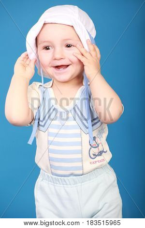 Portrait of a little boy in a bright sailor suit standing there smiling holding hat in hands . Blue background