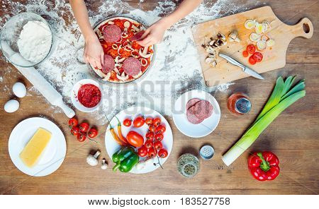 Top View Of Child Making Pizza With Pizza Ingredients, Tomatoes, Salami And Mushrooms On Wooden Tabl
