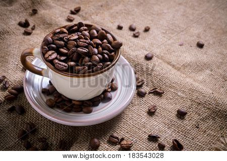 White Cup full of roasted coffee beans on rustic old cloth.