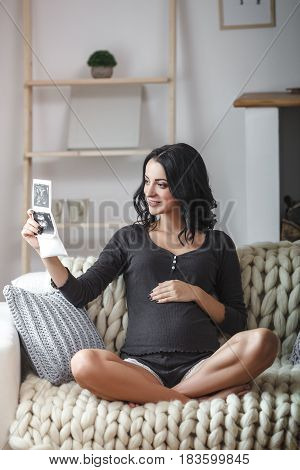happy beautiful bruntte pregnant woman relaxing on sofa with echo scan in hands against window