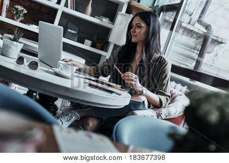 Concentraiting on project. Beautiful young woman holding a pencil and using laptop while sitting in restaurant