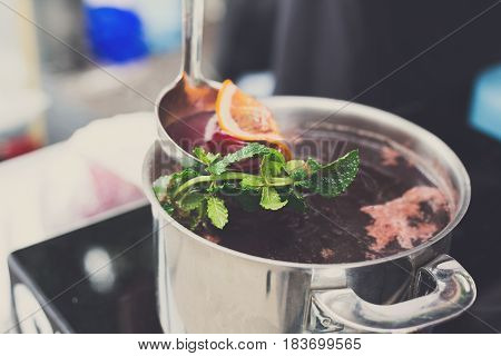 Making mulled wine for sale at country fair. Glintwine with spices, greens, citrus and cloves in big metal stock pot, mixed with soup ladle.