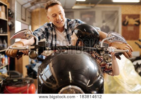 Do you like it. Adorable vibrant pretty girl and her dad having fun at dads workshop while he sitting her on his vehicle and acting like taking a ride