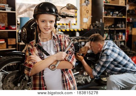 Future expert. Attentive smart committed child holding a wrench and wearing a helmet while helping her dad at his workshop