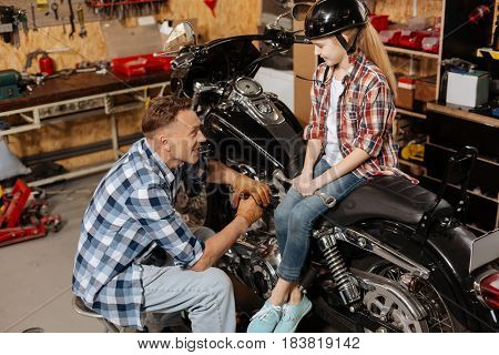 My little advisor. Artistic enthusiastic gorgeous girl sitting on a bike and looking at her father while he fixing a bolt on his motorcycle