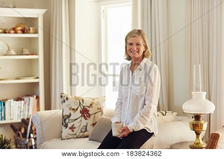 Senior woman looking relaxed and happy in her lounge at home, sitting on the edge of the  arm of her couch holding a coffee cup