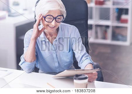 Feeling happiness. Delighted pensioner worker leaning on her right hand looking attentively at folder while sitting on big chair