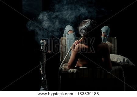 pretty woman with sexy back and body in jeans sitting on eastern chair with shisha bong or kalian as arabic tradition girl smoking hookah on black background