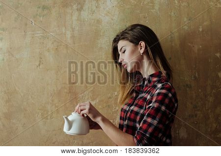 Teatime and ceremony. Pretty girl or beautiful woman with stylish blond long hair in red plaid shirt holding white ceramic tea pot teapot on beige wall