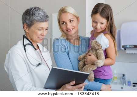 Doctor showing medical report to mother on a digital tablet in hospital. Mother carrying daughter looking at pediatrician holding tablet. Little girl with teddy bear looking at digital tablet screen.