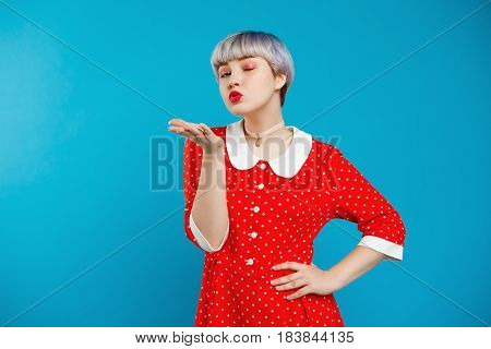 Close up portrait beautiful dollish girl with short light violet hair wearing red dress sending kiss over blue background. Copy space.