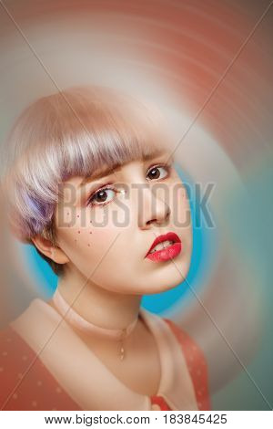 Close up artistic conceptual portrait of beautiful dollish girl with short light violet hair wearing red dress over blue background. Blurry forefront. Copy space.