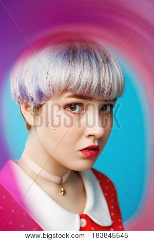 Close up artistic conceptual portrait of beautiful dollish girl with short light violet hair wearing red dress over blue background. Blurry forefront. Copy space.