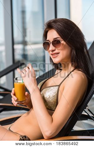 Young attractive slim girl in bikini relaxing on deck chair in wellness spa hotel resort. Woman holding a glass of orange juice