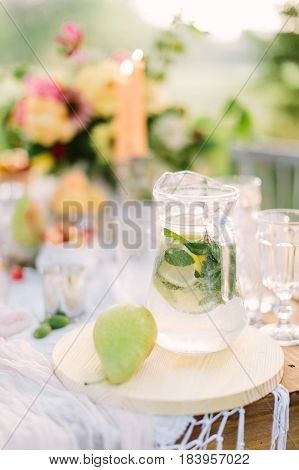 picnic, food, summer, holiday concept - glass jug full of lemonade on festive table, pear, flagon with water, slices of lemon, lime, mint, background of bouquet, candles, glasses