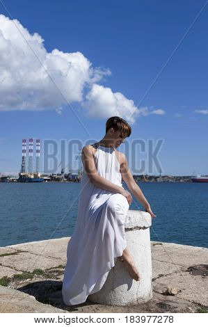 Female sitting on pier in white dress with blue skies and sea in background.