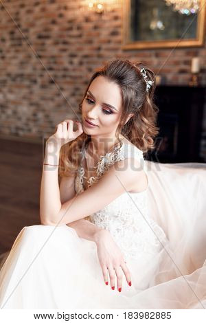 Wedding. Bride in beautiful dress sitting on sofa indoors in studio interior like at home. Trendy wedding style shot in full length. Young attractive caucasian brunette model like a bride