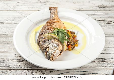 Dorada Stuffed With Vegetables. White Plate With Grilled Dorada Fish On A Wooden Table Close Up And 