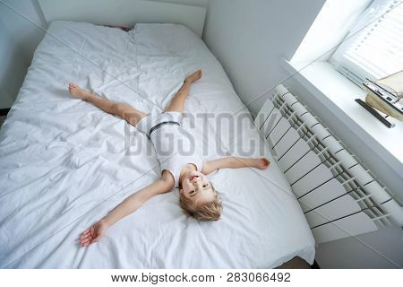 Happy Boy Playing In White Bedroom. Little Boy Brother Play On The Bed Wearing Pajamas. Family At Ho