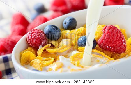 Pouring Milk Into Bowl Of Corn Flakes With Berries. Ready-to-eat Breakfast Of Cornflakes With Raspbe