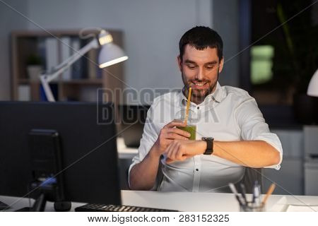 business, technology and time management concept - happy smiling businessman drinking green smoothie and looking at smart watch at nigh office