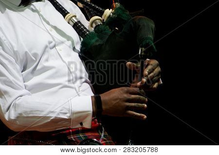 A Black Man In A White Shirt Is Holding A Scottish Bagpipe. Multicultural On A Black Background.