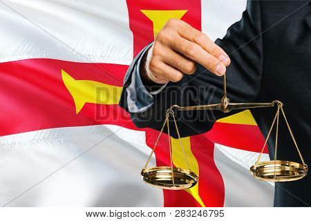 Judge Is Holding Golden Scales Of Justice With Guernsey Waving Flag Background. Equality Theme And L