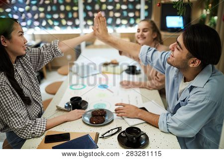 Cheerful Teenage Students Giving High Five To One Another Over Table With Cups Of Tea And Other Stuf