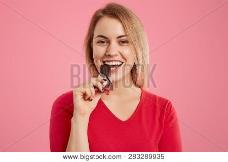Photo Of Attractive Light Haired European Young Woman Eats Delicious Sweet Chocolate, Being Sweettoo
