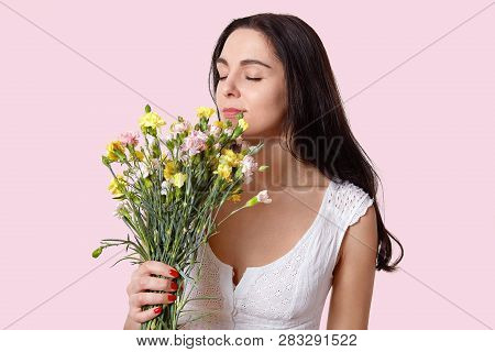 Horizontal Shot Of Pleasant Looking Caucasian Woman With Black Hair, Closes Eyes With Pleasure, Smel