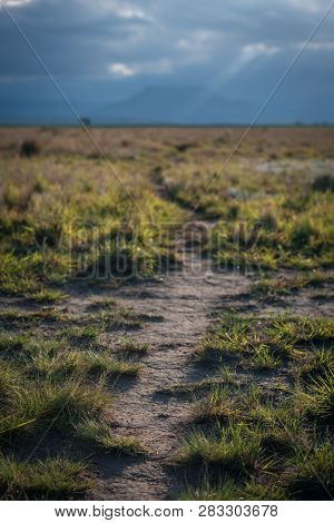 A Hiking Trail Leads Across A Grassy Plain Towards The Hazy Mountains In The Distance, Lit Up By The