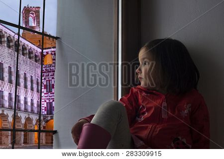 Little Girl Observing The Badajoz Plaza Alta Sitting In Her Home Sill. She Is Wearing Raincoat
